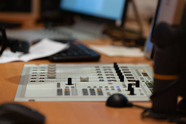 A detailed view of an audio mixing console on a wooden desk, showcasing various faders and buttons. The workspace is cluttered, hinting at a busy production setting.