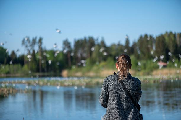 A woman in her 30s stands by a serene lake watching birds fly over the water, with a lush forest in the background.