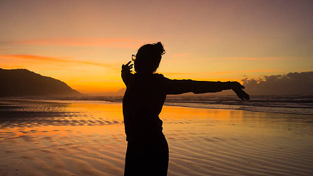 Experience a moment of tranquility and renewal with this stunning image. A lone figure is captured in silhouette, performing stretching exercises on a serene beach as the first light of day breaks. The sky is painted in breathtaking shades of orange and red, reflecting beautifully on the calm sea waters at the shoreline. The strong backlight emphasizes the person's form against the vibrant backdrop, creating a powerful visual of focus, health, and connection with nature. This photograph evokes themes of wellness, mindfulness, exercise, new beginnings, solitude, and the beauty of the natural world at dawn. Perfect for wellness blogs, travel websites, motivational content, or health-related campaigns.
