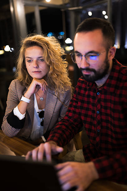 Two colleagues collaborating on a project at a laptop in a cozy indoor setting, highlighting teamwork and focus. The ambiance suggests a night working environment filled with concentration and partnership.