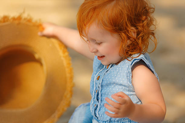 Cheerful red-haired toddler girl in a blue dress smiling and playing with her hands outdoors on a sunny summer day. Bright natural light, warm tones, and joyful expression .