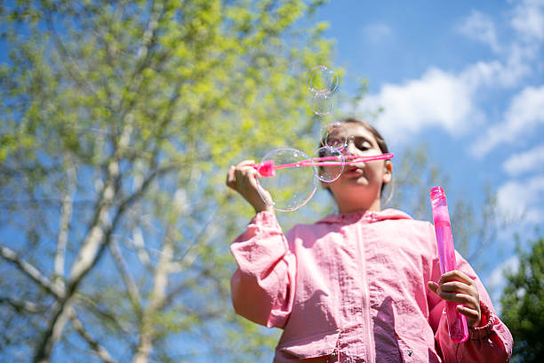 A young girl is blowing colorful soap bubbles outdoors against a vibrant, sunny backdrop with blue sky and greenery. The scene captures the playful spirit of childhood and the beauty of nature.