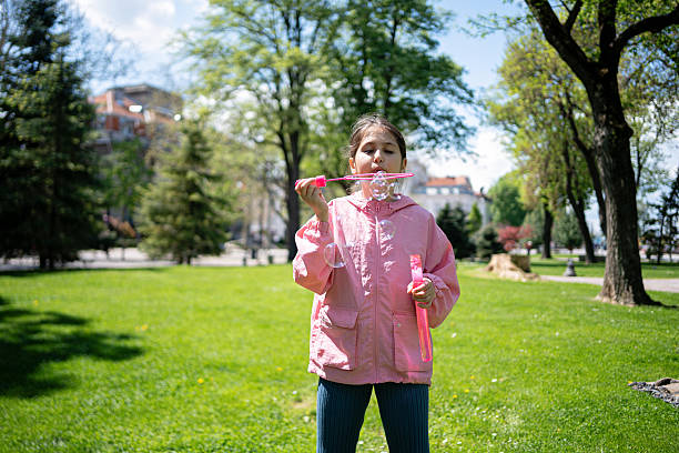 A child enjoys a playful moment blowing soap bubbles amidst a lush green park under daylight. The sunny weather and outdoor environment emphasize the carefree and joyful nature of the activity.
