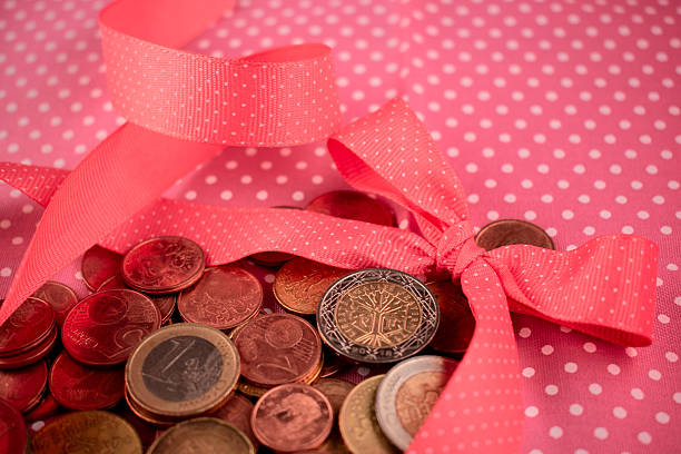 A pile of assorted coins lies beneath a neatly tied pink ribbon with white polka dots. The coins, including copper and a notable two-euro piece, form a bed beneath the bow, suggesting concepts of financial gifts or savings. The ribbon adds a festive or celebratory touch, with a background hinting at a vibrant, colorful setting. The arrangement of elements may symbolize budgeting, gift-giving, or investments.