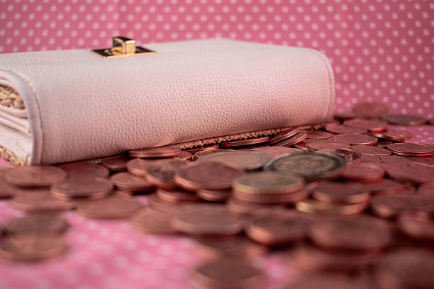 A white leather wallet with a gold clasp is laid on a pink polka dot fabric. Scattered around it are various euro coins, including cents and 1 and 2 euro coins. The wallet's texture appears smooth, while the coins contrast with metallic tones. The arrangement suggests themes of finance, saving, and monetary management.