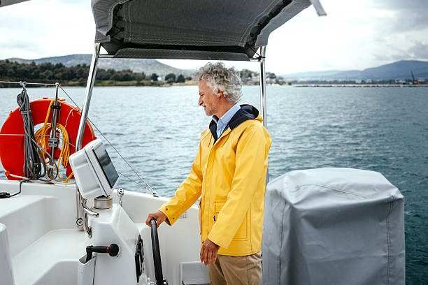 A senior boat captain wearing yellow raincoat steering a catamaran