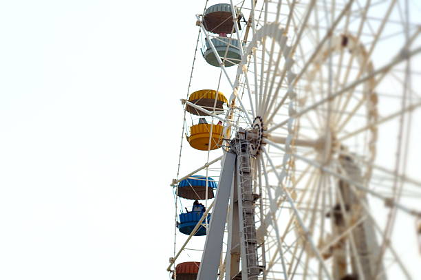 Close-up view of ferris wheel passenger gondolas in vibrant colors including red, yellow, blue and green arranged along the wheel structure against overcast sky with bare winter trees in background. The composition emphasizes the cheerful carnival colors and mechanical details of the amusement ride while soft natural lighting creates pleasant atmosphere despite the winter season. Multiple gondolas visible at different positions showcase the scale and engineering of the fairground attraction with white structural framework supporting the colorful passenger cars. This festive carnival scene embodies themes of amusement parks, family entertainment, seasonal fairs, childhood joy, and recreational activities. The bright colors and nostalgic appeal create strong visual appeal perfect for family entertainment marketing, carnival promotions, tourism advertising, childhood memory content, or recreational activity guides. The image commercially valuable for amusement park operators, fair organizers, family entertainment venues, or wellness content about the importance of play and recreational activities that bring families together and create lasting memories through shared experiences at seasonal celebrations and community events.​​​​​​​​​​​​​​​​