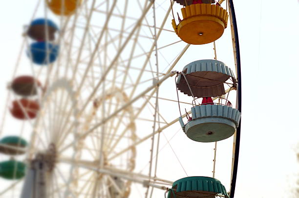 Upward perspective of ferris wheel showing colorful passenger gondolas in orange, blue, brown and green suspended from white steel framework against overcast sky. The composition captures the engineering details and vibrant carnival atmosphere with multiple gondolas visible at different heights while structural spokes and support cables create geometric patterns. Soft natural lighting emphasizes the cheerful colors and mechanical precision of the amusement ride creating nostalgic fairground ambiance. This classic carnival scene embodies themes of amusement parks, family entertainment, fairground attractions, childhood memories, and recreational joy. The nostalgic appeal and bright colors create strong visual appeal perfect for carnival marketing, family entertainment promotions, tourism advertising, amusement park guides, or childhood nostalgia content. The image commercially valuable for fairground operators, amusement park marketing, family entertainment venues, or wellness content about the importance of recreational activities and shared family experiences that create lasting memories through traditional carnival attractions that bring communities together for seasonal celebrations and provide multigenerational entertainment opportunities.​​​​​​​​​​​​​​​​