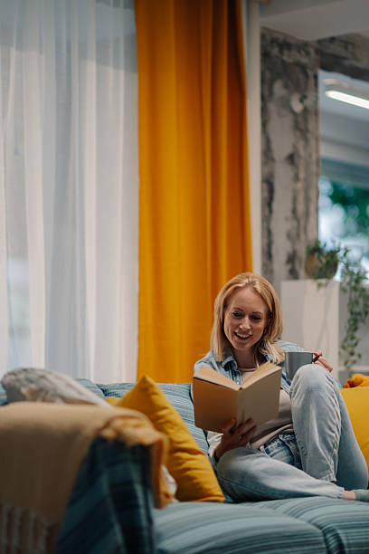 istock Smiling woman reading a book and drinking tea on the sofa at home 2216761669