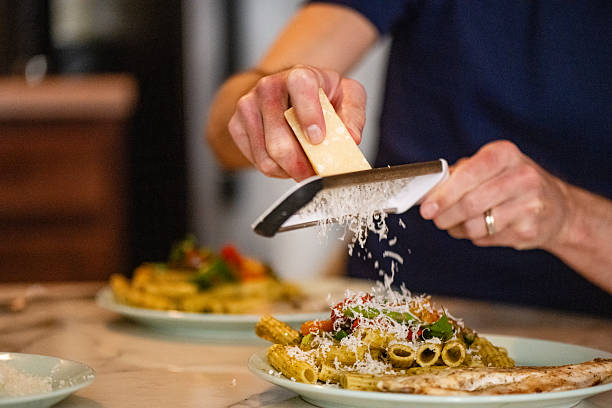 This is a close up photograph of the hands of a gay white man in his 30s hand grating a block of Parmesan cheese over a homemade pasta dinner.