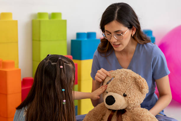 Latin american pediatrician uses a teddy bear to explain medical procedures to a preschool age girl in a colorful doctor's office