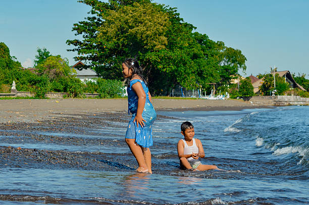 The girl walks on the wet sand while the boy remains seated, playing in the gentle surf.