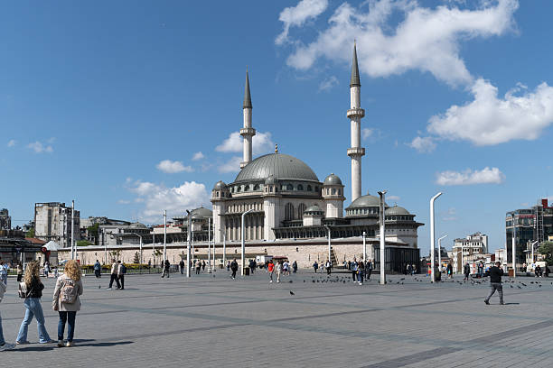Istanbul, Turkey - May 25 ,2025: A wide-angle view capturing the expansive Taksim Square in Istanbul, Turkey, prominently featuring the modern Taksim Mosque with its distinctive minarets and dome. The square is bustling with pedestrians, including people walking with their dogs, under a bright blue sky with scattered clouds. This image conveys the dynamic atmosphere of one of Istanbul's most central and significant public spaces.