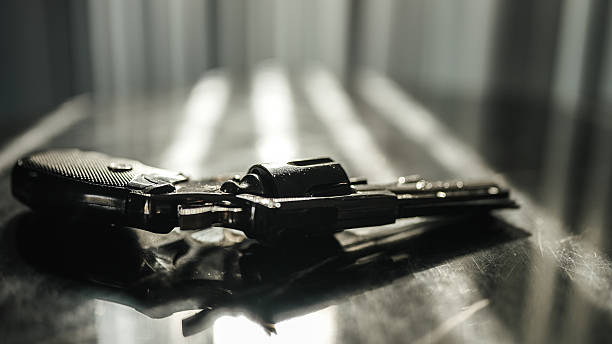 istock Shiny black revolver on a black table backlight by dramatic sunlight through the window blinds. Cinematic crime scene suicide concept. 2216789255