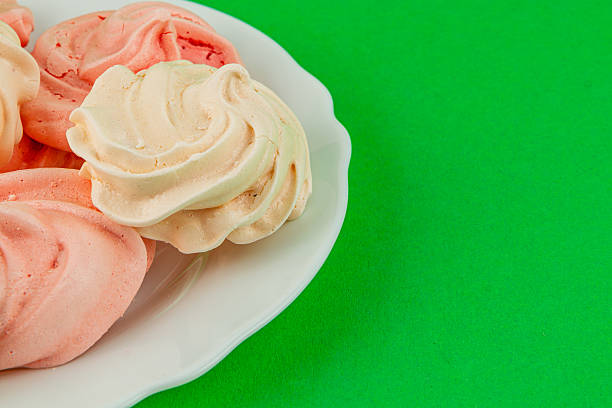 A selection of pink and white meringue cookies sits on a white plate against a vibrant green backdrop, showcasing their delicate texture and swirl design.