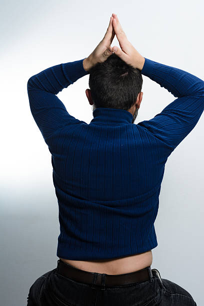 A rear view of a man wearing a blue turtleneck shirt and dark pants. His hands are joined together above his head against a plain backdrop.