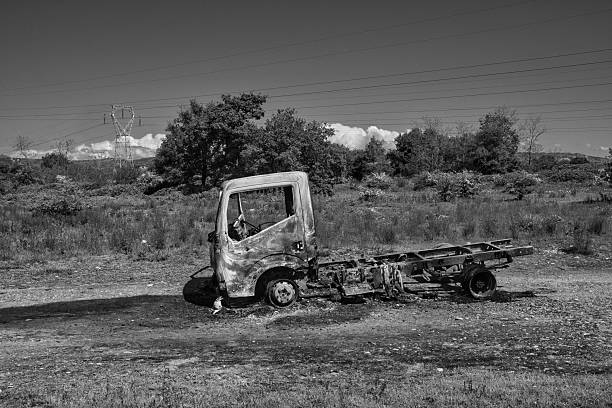 Industrial and domestic waste in nature. Visual composition of landscape with a burnt truck. Only the steel structure is visible. The ground is blackened. The landscape of trees and the clear sky contrast sharply.
