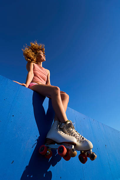 Young woman sits on a blue ledge, wearing roller skates, basking in the sunlight. Her curly hair flows in the breeze against a clear blue sky