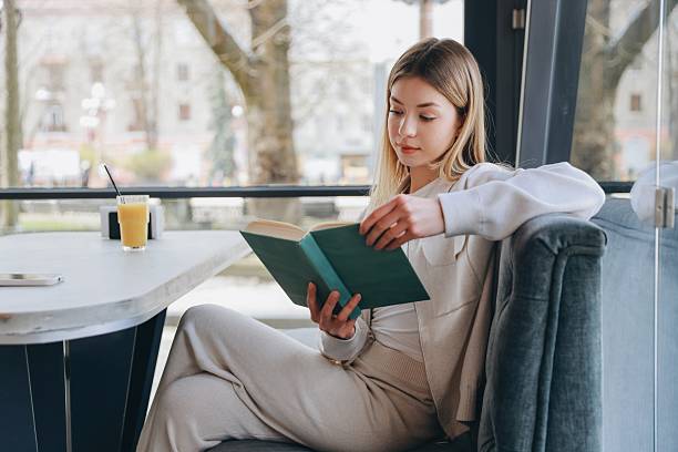 Young woman enjoying a captivating book while sipping a refreshing drink in a bright, cozy cafe, surrounded by natural light and tranquility