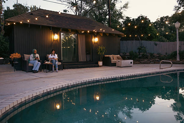 A modern outdoor patio with a wooden deck, featuring a dining table and chairs under a sleek roof. A swimming pool is visible in the foreground, illuminated by soft lighting at dusk.