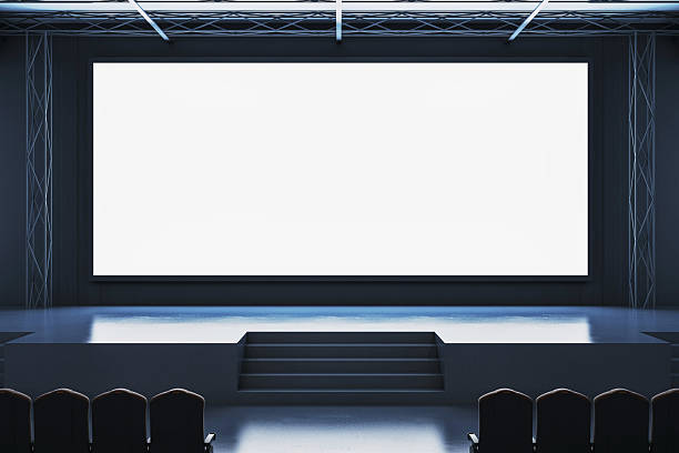 istock Large empty white display screen on modern conference stage with dark interior lighting and chairs in front in corporate hall setting. 3D Rendering 2216829333