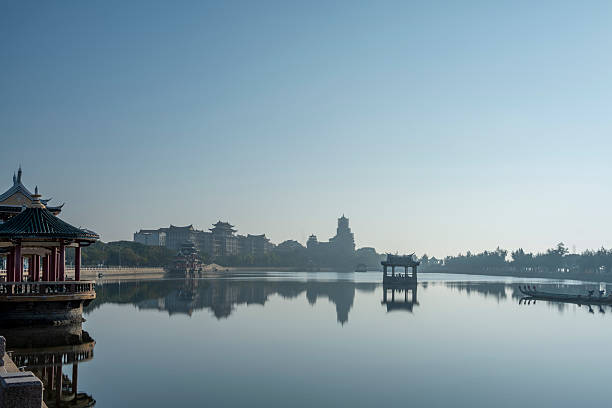 A serene lakeside view featuring traditional Chinese architecture, including a pagoda and pavilion, set against a clear blue sky. The reflection on the water adds to the peaceful atmosphere. Ideal for travel or cultural themes.