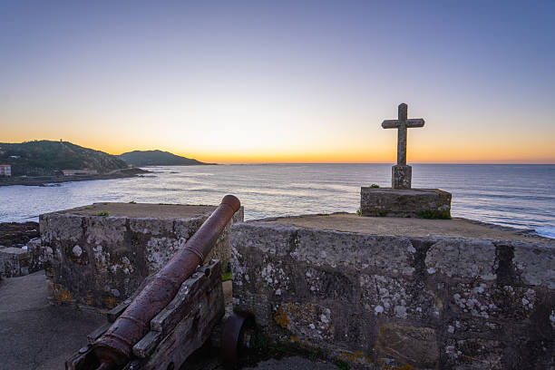 A historic defensive cannon and a stone cross stand guard over the beautiful Baiona Bay, Spain, at sunset.