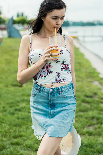 A girl is standing by the lake with a glass of juice in her hands. A girl in a T-shirt and skirt by the lake. The girl is holding a hat in her hands.