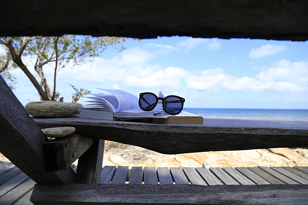 A man enjoys a quiet moment on a sunbed by the sea, engrossed in a book while the clear blue sky and mountains create a stunning natural backdrop.
