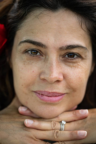 Intimate and detailed portrait of a middle-aged woman with natural skin and a soft, genuine expression. Her hands gently support her chin, and the close framing draws attention to her eyes and face, capturing the essence of real beauty, confidence, and warmth.