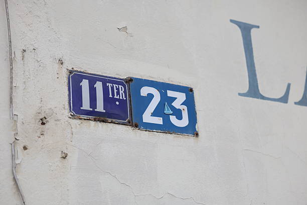 Two street number signs are affixed to a white wall with visible cracks and peeling paint. The sign on the left reads 11 TER in white text on a blue background, while the sign on the right displays 23 with a small sailboat icon between the numbers.