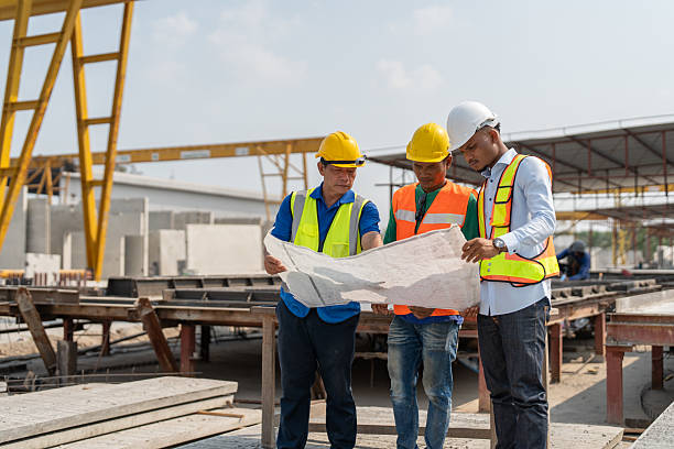 istock Senior Asian foreman and worker wearing protective helmets and vests explaining on building blueprint in construction site 2216939522