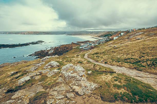 A hillside with a house and a lighthouse on top in England. The house is white and the lighthouse is tall. The hillside is lush and green, and the ocean is in the background. The scene is peaceful and serene