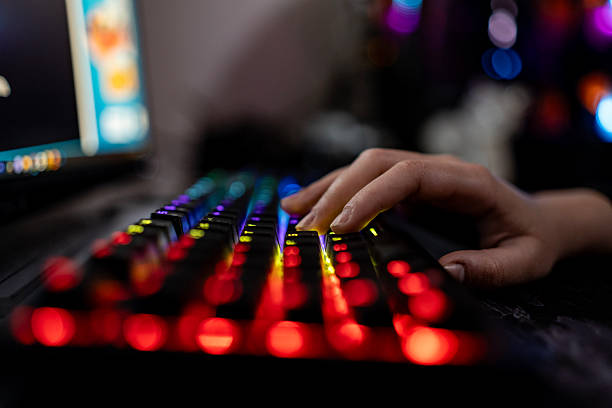 A hand typing on a vibrant RGB backlit mechanical keyboard with a blurred monitor in the background, creating a visually captivating atmosphere in low-light conditions.