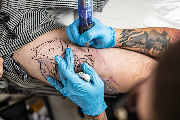 Close-up of a tattoo artist wearing gloves while working on a client's leg tattoo design in a studio.