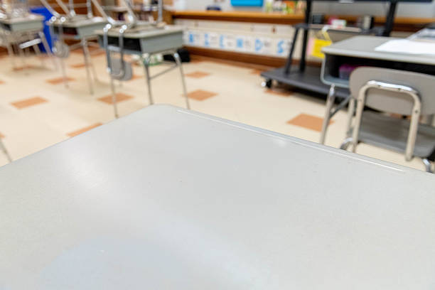 Wide view of empty elementary school classroom with student desks and chairs arranged for learning, with natural lighting creating welcoming educational environment and floor tiles shown in the background.