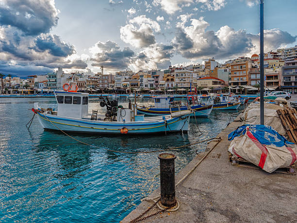 Colorful fishing boats bob gently in the harbor of Sitia, Greece, under a dramatic sky filled with clouds.