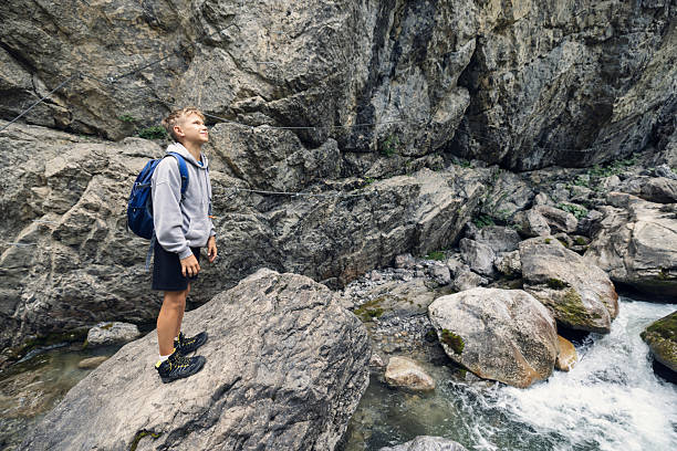 A teenage boy hiker, dressed in a grey hoodie, black shorts, and sturdy hiking shoes, stands on a large rock amid a rugged natural landscape. With a blue backpack strapped securely to his shoulders, he is smiling and admiring the surroundings. The terrain is rocky and uneven, with a small stream carving through the wilderness. Towering rock formations in the background suggest a remote and adventurous location, possibly deep within a mountainous or forested region.\nShot with Canon R5 on a summer day.