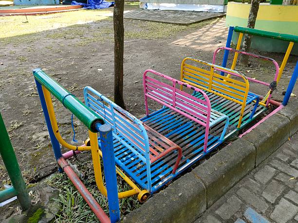 a colorful metal seesaw designed for children, located in an outdoor playground area. The seesaw has multiple seats painted in vibrant colors such as pink, yellow, blue, and green. It appears slightly worn from use but still functional. The surrounding area includes paved paths, dirt, and some grass, indicating an open recreational space.