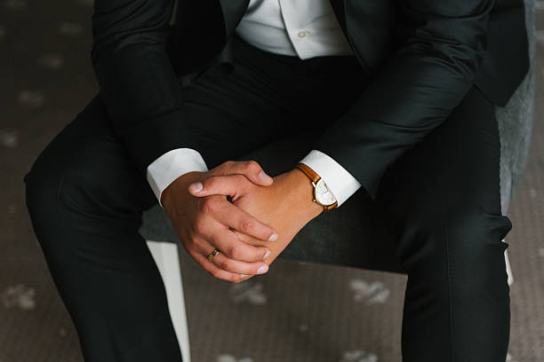 A close up of the groom sitting in a black suit, wearing a classic watch, hands folded in a relaxed yet elegant pose.