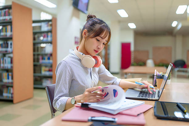 istock Young Asian Woman Studying with Laptop in Library Bookshelves Learning Education Knowledge Research Information Data Analysis Charts Graphs and Headphones 2216977892