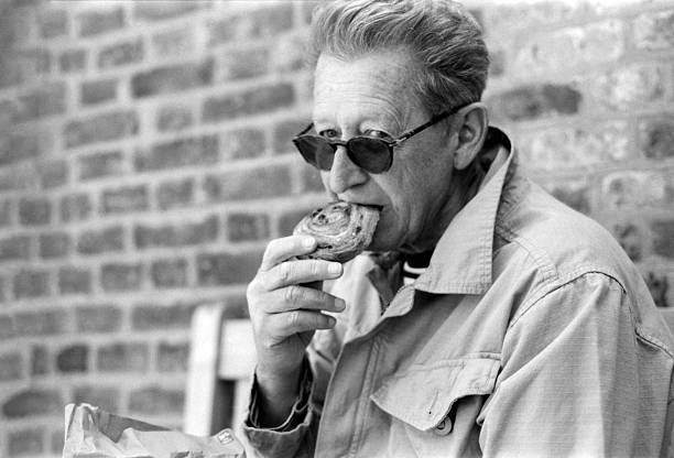 Grainy monochrome analog photo capturing a senior man wearing sunglasses, savoring a delicious pastry while leaning against a textured brick wall, radiating contentment and relaxation