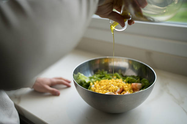 Professional chef is carefully pouring olive oil over a colorful salad with corn, lettuce, and tomatoes in a stainless steel bowl, preparing a healthy and delicious meal