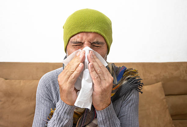Mature man with a cold sneezing into tissue, wrapped in blanket, wearing beanie and scarf, resting on couch. Concept of flu, illness, winter, home care.