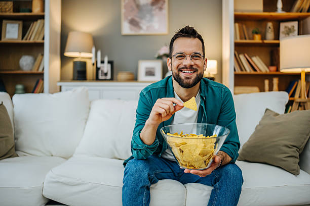 istock Young man sitting on sofa at home eating nachos and smiling 2216997087