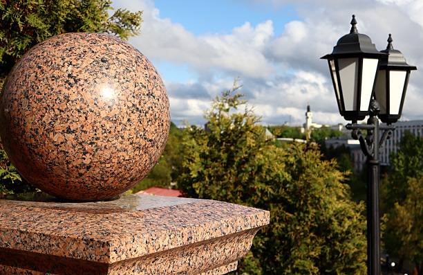 A polished granite sphere decorates a stone post in an urban park. A vintage-style street lamp and green trees complete the elegant architectural setting.