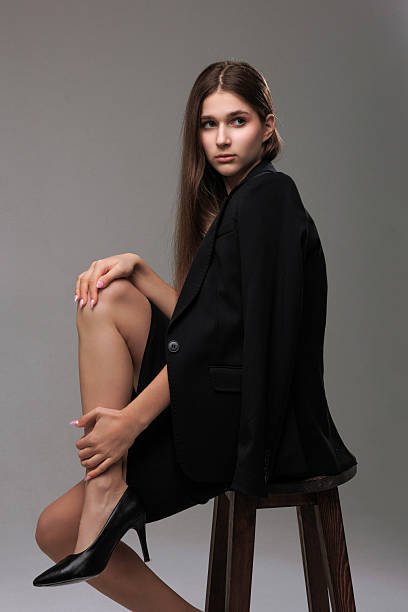 A young model is seated on a wooden stool, showcasing a poised expression. She wears a chic black blazer and polished heels, highlighted against a soft, neutral background.