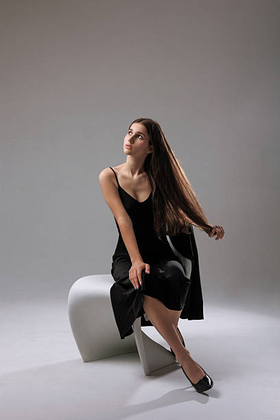 A model showcases a stylish black dress while sitting gracefully on a modern stool in a well-lit studio. Her long hair flows elegantly, creating a striking look.