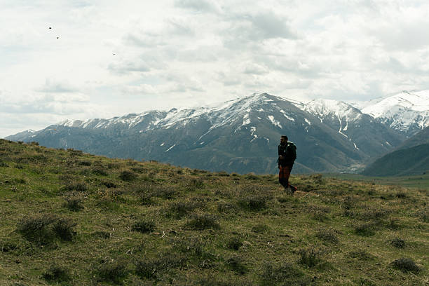 Hiker walks on a grassy mountainside with snowy peaks in the background, enjoying the tranquility of nature
