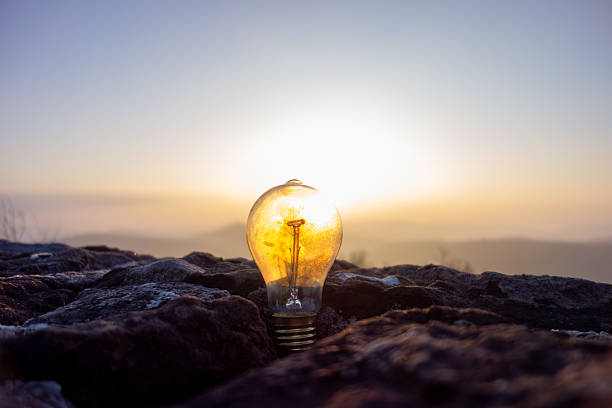 A light bulb rests on a rocky surface as the sun rises in the background, illuminating the landscape with warm golden hues. The contrast of nature and technology is inspiring.